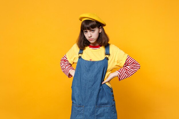 Portrait of serious girl teenager in french beret, denim sundress standing with arms akimbo on waist isolated on yellow wall background. People sincere emotions, lifestyle concept. Mock up copy space.