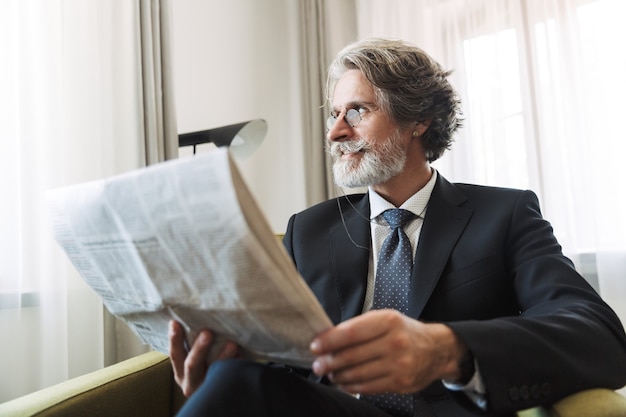 Portrait of a serious grey-haired senior businessman indoors at home dressed in formal clothes reading newspaper.