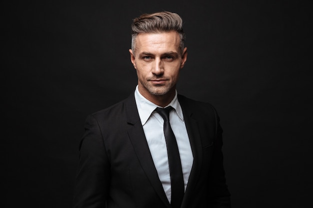 Portrait of serious handsome businessman dressed in formal suit posing and looking at camera isolated over black wall