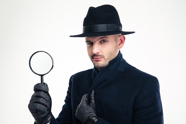 Photo portrait of serious handsome detective in black coat, hat and gloves holding magnifying glass over white wall