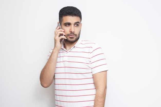 Portrait of serious handsome young adult man wearing T-shirt standing, using smartphone, making call and talking with friend. Indoor, isolated, studio shot, white background