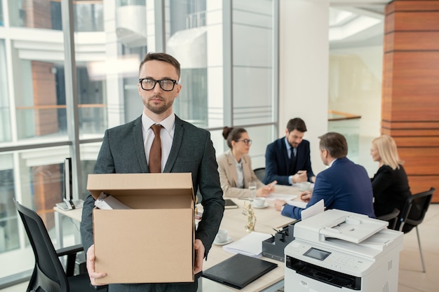 Portrait of serious handsome young lawyer in formal suit and eyeglasses standing with box of stuff, he leaving office after dismissal