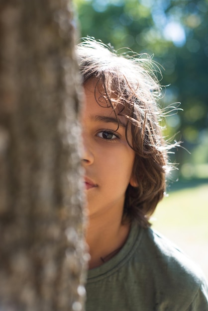 Portrait of serious little boy in park. Dark-haired boy peeking out from tree, looking at camera. Family, love, childhood concept