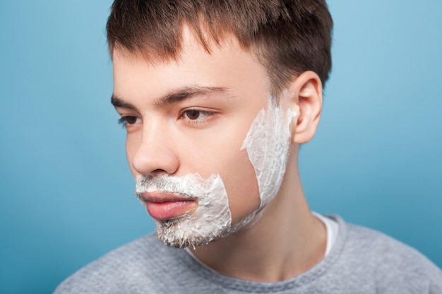 Portrait of serious man in casual clothes looking aside with shaving foam on his bristle and mustache showing smooth soft skin on shaved part of cheek indoor studio shot isolated on blue background