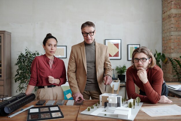 Portrait of serious modern architects standing at desk with 3D model of house while working with building maquette