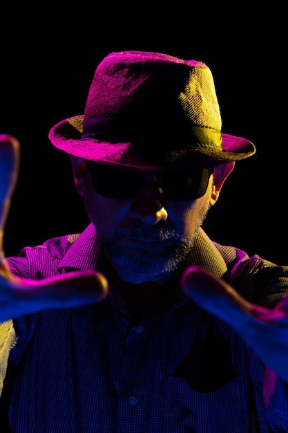 portrait of a serious mysterious man with a beard wearing a hat and sunglasses making gestures with his hands portrait in studio with colored lights and dark background