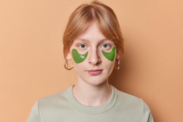 Portrait of serious red haired millennial woman with freckled skin applies green collagen patches under eyes looks confident at camera wearrs earrings and t shirt isolated over brown background