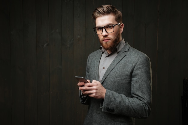 Portrait of a serious smart man in casual suit and eyeglasses holding mobile phone