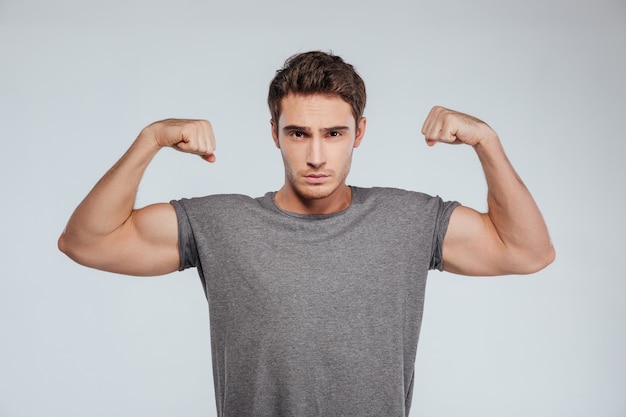 Portrait of a serious strong man flexing both biceps and looking at camera on the gray