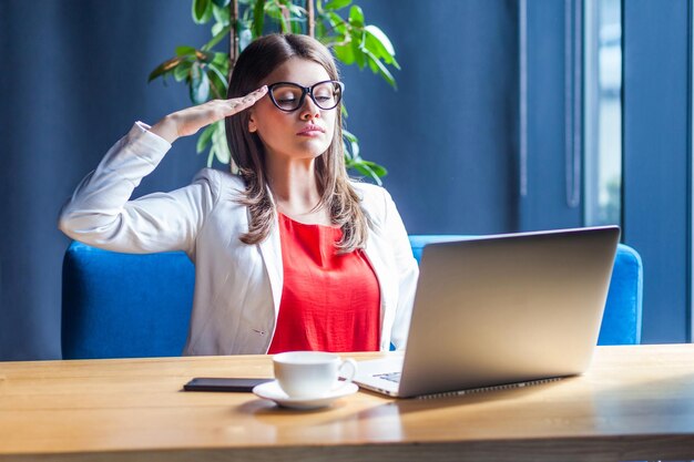 Portrait of serious stylish brunette young woman in glasses\
sitting looking at her laptop screen on video call and giving\
salute on boss command indoor studio shot cafe office\
background