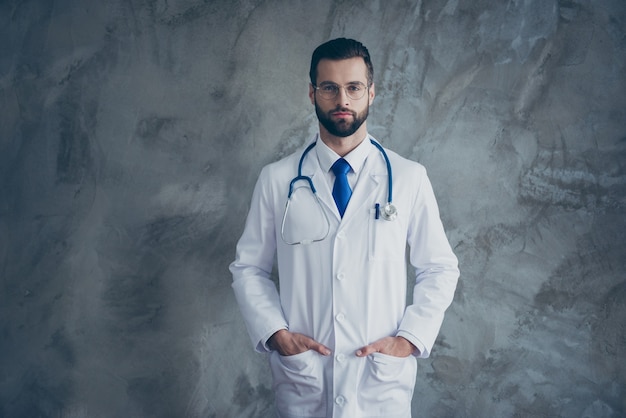 Portrait of serious talented doctor put his hands in pockets of white coat look ready to cure patients in clinic isolated over grey color wall