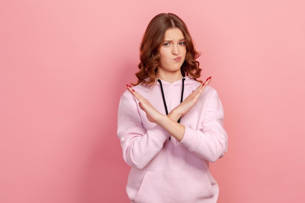 Портрет серьезной девушки-подростка с каштановыми волосами в толстовке с капюшоном, делающей знак x со скрещенными руками, жестикулируя стоп-предупреждением о запрете языка тела. Снимок в помещении на розовом фоне