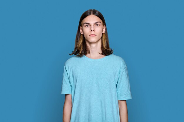 Portrait of serious teenage guy looking at camera on blue color background handsome young male with long hair hairstyle posing in studio youth students people concept