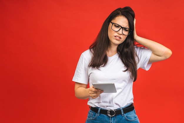 Portrait of a serious tired unhappy young brunette business woman using tablet computer isolated over red background