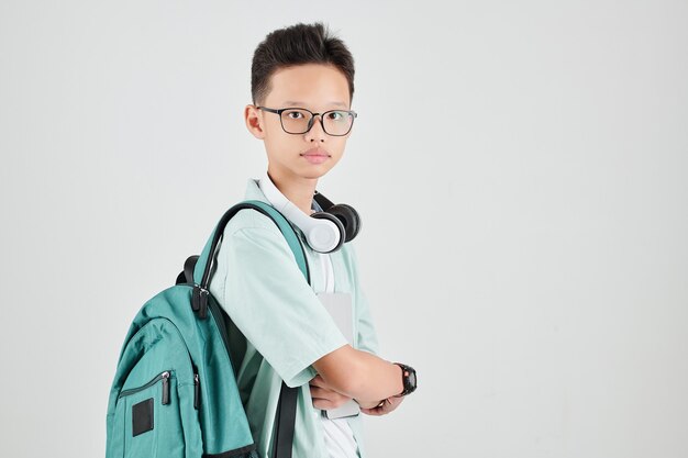 Portrait of serious Vietnamese schoolboy in glasses posing with backpack, headphones and tablet computer