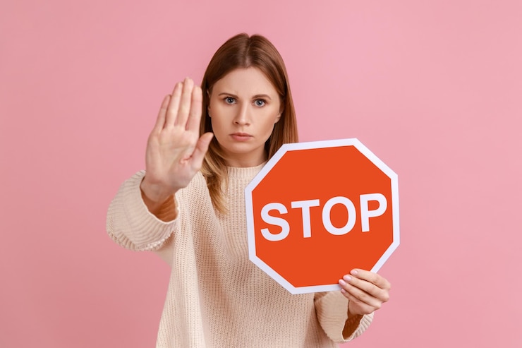 Photo portrait of serious woman holding red stop sign, looking at camera with ban palm gesture, has strict expression, prohibition, wearing white sweater. indoor studio shot isolated on pink background.