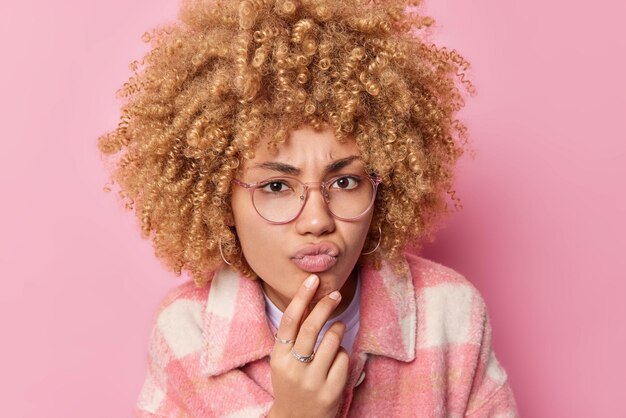 Foto il ritratto di una donna seria con le labbra delle borse dei capelli ricci ha uno sguardo attento guarda qualcosa che indossa gli occhiali e la giacca ascolta le pose di informazioni su sfondo rosa espressioni del viso umano