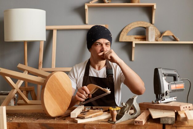 Portrait of serious young adult man carpenter wearing white tshirt black cap and brown apron sanding wood block looking at camera keeps finger under hose