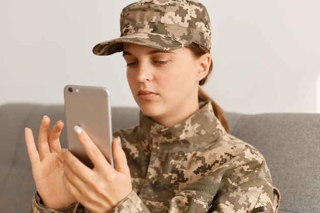 Ritratto di un soldato militare adulto giovane serio che indossa un'uniforme mimetica e un cappuccio che tiene il telefono cellulare e digita i messaggi di risposta o controlla la posta elettronica