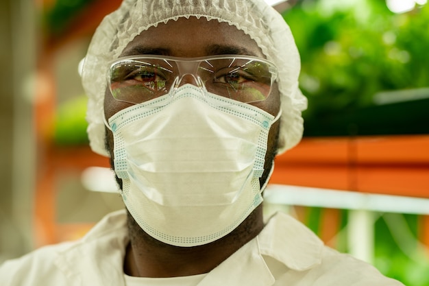 Портрет серьезного молодого афроамериканского специалиста по выращиванию в маске, очках и кепке, стоящего на вертикальной полке фермы