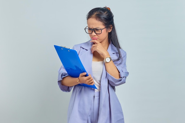 Portrait of serious young Asian business woman holding chin, contemplating something while holding document folder isolated on purple background