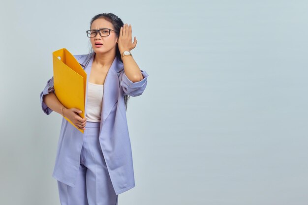 Portrait of serious young Asian business woman keeps hand near ear tries to overhear private conversation listens gossips while holding yellow folder isolated on purple background