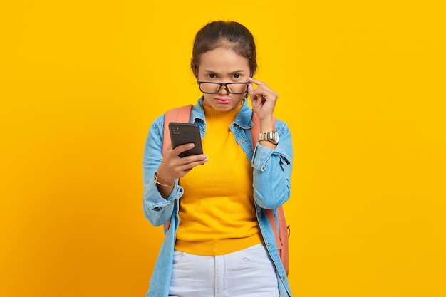 Portrait of serious young asian woman student in casual clothes\
with backpack using smartphone and taking off glasses isolated on\
yellow background. education in college university concept