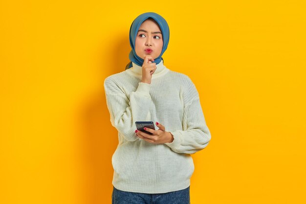 Portrait of serious young asian woman in white sweater and hijab thinking about question with hand on chin while holding mobile phone isolated over yellow background People islam religious concept