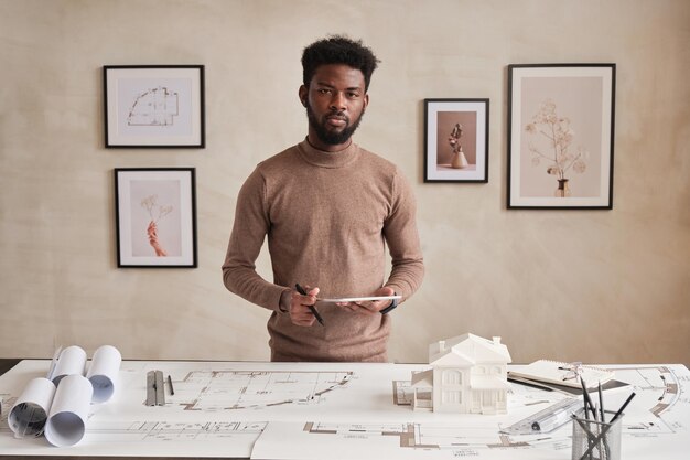 Portrait of serious young bearded black architect in sweater standing with tablet at table full of sketches and supplies in contemporary office