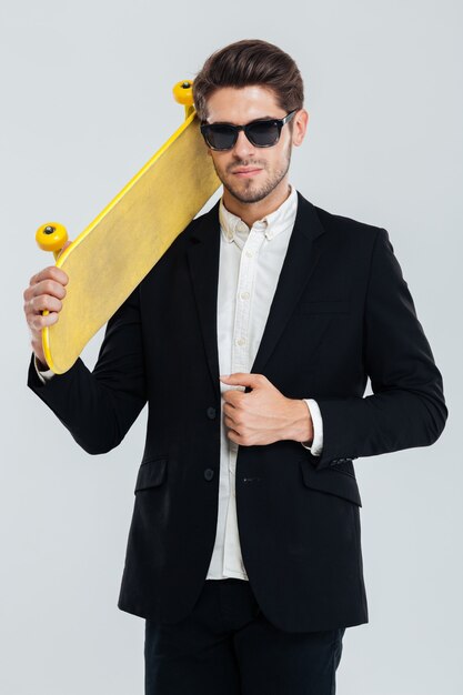 Portrait of serious young businessman in sunglasses holding yellow skateboard on his shoulder over gray wall