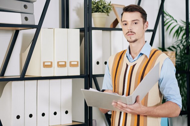 Portrait of serious young businessman with moustache and beard searching for document in archive in office