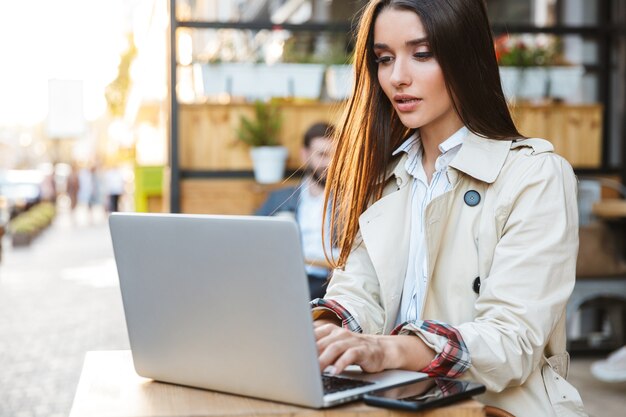 Портрет серьезной молодой бизнес-леди в формальной одежде, сосредоточенной во время работы на ноутбуке в кафе на открытом воздухе