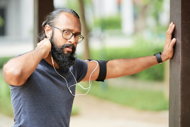 Portrait of serious young Indian runner with beard listening to music in earphones while training outdoors