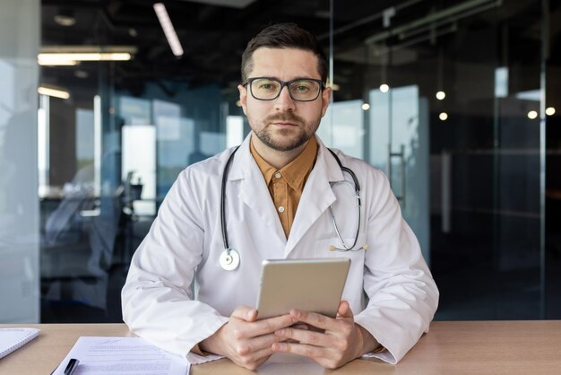 Portrait of a serious young male doctor sitting in the office with a tablet in his hands in a white coat and talking on a video call with a patient Professional online conference