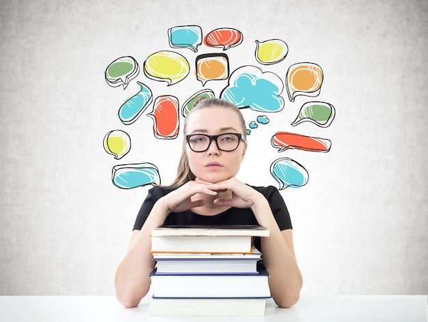 Portrait of a serious young woman in glasses looking forward and sitting with her chin on a stack of books. A concrete wall background with colorful speech bubbles.