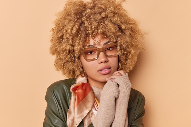 Portrait of serious young woman has blonde curly hair healthy skin looks directly at camera with calm expression wears stylish clothes transparent eyeglasses poses against beige studio wall.