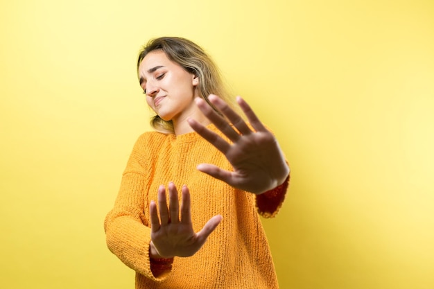 Photo portrait of a serious young woman showing stop gesture with her palm over yellow background