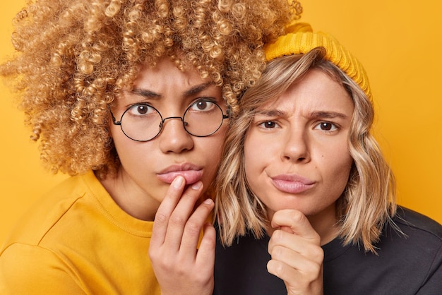 Portrait of serious young women focused attentively at camera\
observe something look scrupulously stand next to each other\
dressed casually isolated over yellow background hmm let us\
think