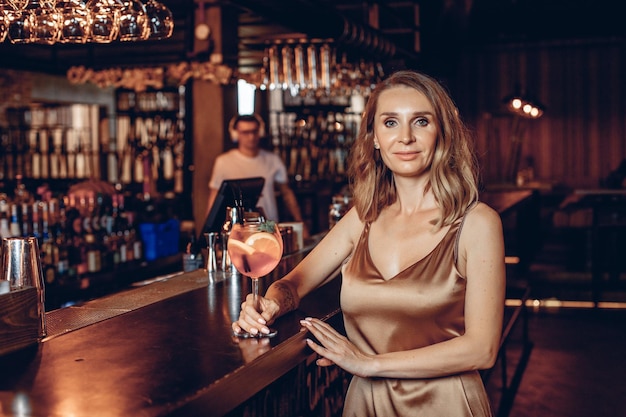 Portrait of sexual attractive lovely young woman wearing golden dress and drinking cocktail at the bar in the club. Party night concept