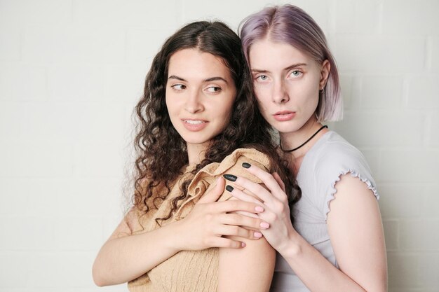 Portrait of sexual woman with purple hair touching shoulders of wavyhaired girlfriend against wall