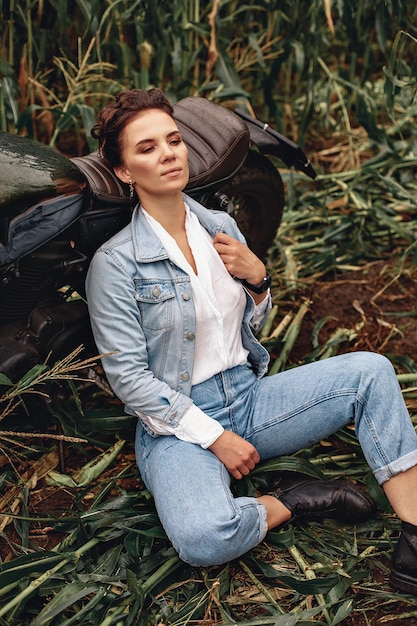 Portrait of a sexy charming biker woman posing and sitting on the ground next to her black retro motorcycle, leaning her back against its seat and looking into the distance. Extreme concept