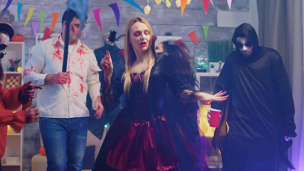Ritratto di sexy incantatrice malvagia che balla alla festa di halloween circondata dai suoi amici in una casa decorata