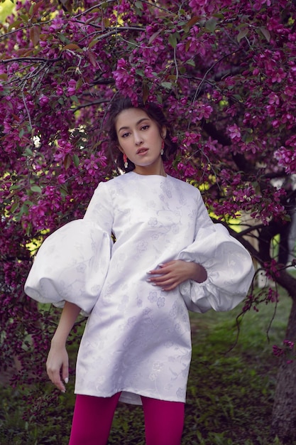 Portrait sexy young woman in a white dress and purple tights stands by the blooming purple cherry trees in spring