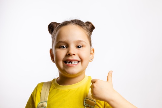 Portrait of shining little girl showing missing front baby tooth and thumb up in yellow tshirt on wh...