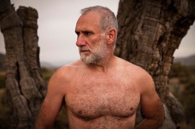 Photo portrait of shirtless adult man standing on bare olive tree against sky