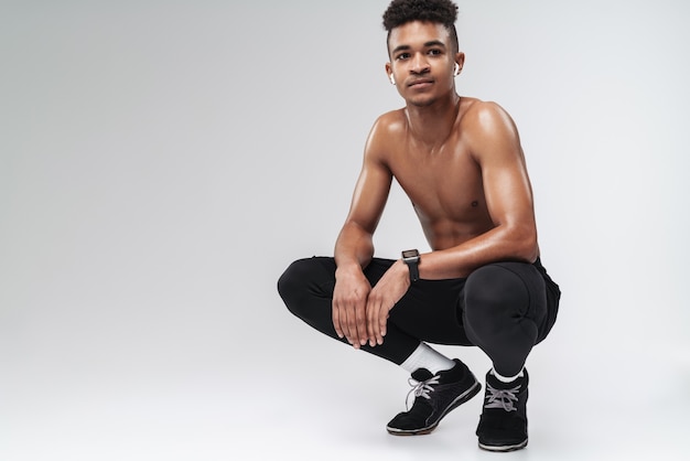 Portrait of shirtless african american man using earpodфs while squatting isolated on grey