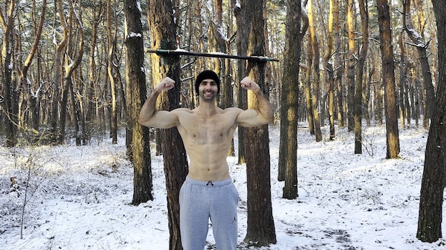 Portrait of shirtless sportsman standing in snowy forest and showing biceps Handsome strong athlete