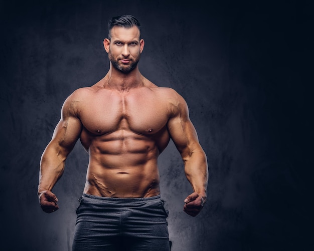 Portrait of a shirtless tall huge male with a muscular body with a stylish haircut and beard, in a sports shorts, standing in a studio. Isolated on a dark background.