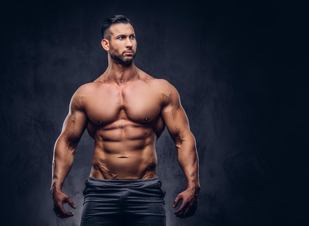 Portrait of a shirtless tall huge male with a muscular body with a stylish haircut and beard, in a sports shorts, standing in a studio. Isolated on a dark background.