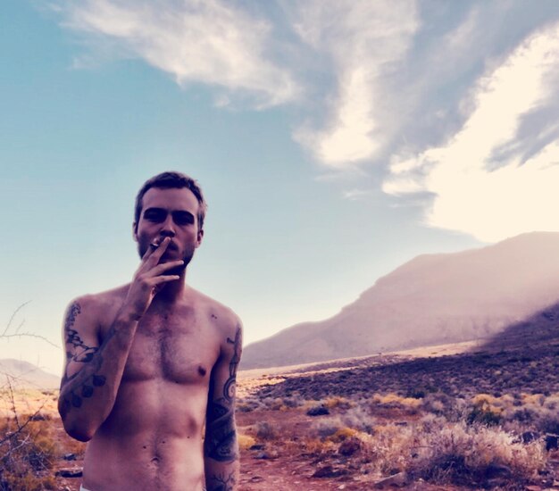 Portrait of shirtless young man smoking on field against sky